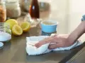 Mano limpiando una encimera con un paño blanco, junto a un frasco de bicarbonato y limones.