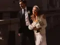 Novia sonriente con velo y ramo, luciendo una chaqueta novia boho sobre su vestido. El novio, con traje oscuro, la abraza.