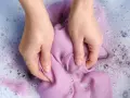 Manos lavando ropa morada a mano en agua con jabón y burbujas.