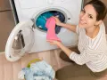 Mujer sonriente carga ropa en lavadora. El detergente sirve para dejar la ropa impecable y eliminar manchas.