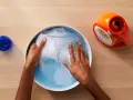 Manos lavando ropa blanca en un bol con agua y detergente. A un lado, un bote de detergente y un tapón azul.
