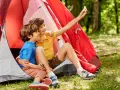Dos niños, uno con talla niño 7 años, se toman un selfie sonrientes dentro de una tienda de campaña roja en un bosque.