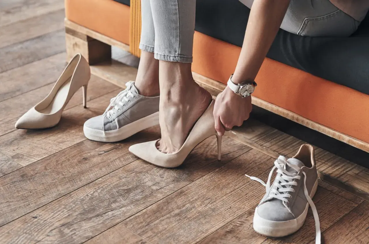 Mujer eligiendo entre varios tipos de pies: zapatillas deportivas y tacones elegantes.