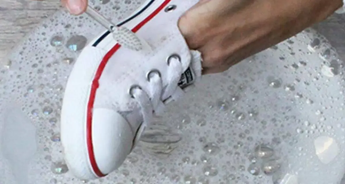 Manos limpian zapatillas de tela de color con cepillo en agua con jabón.
