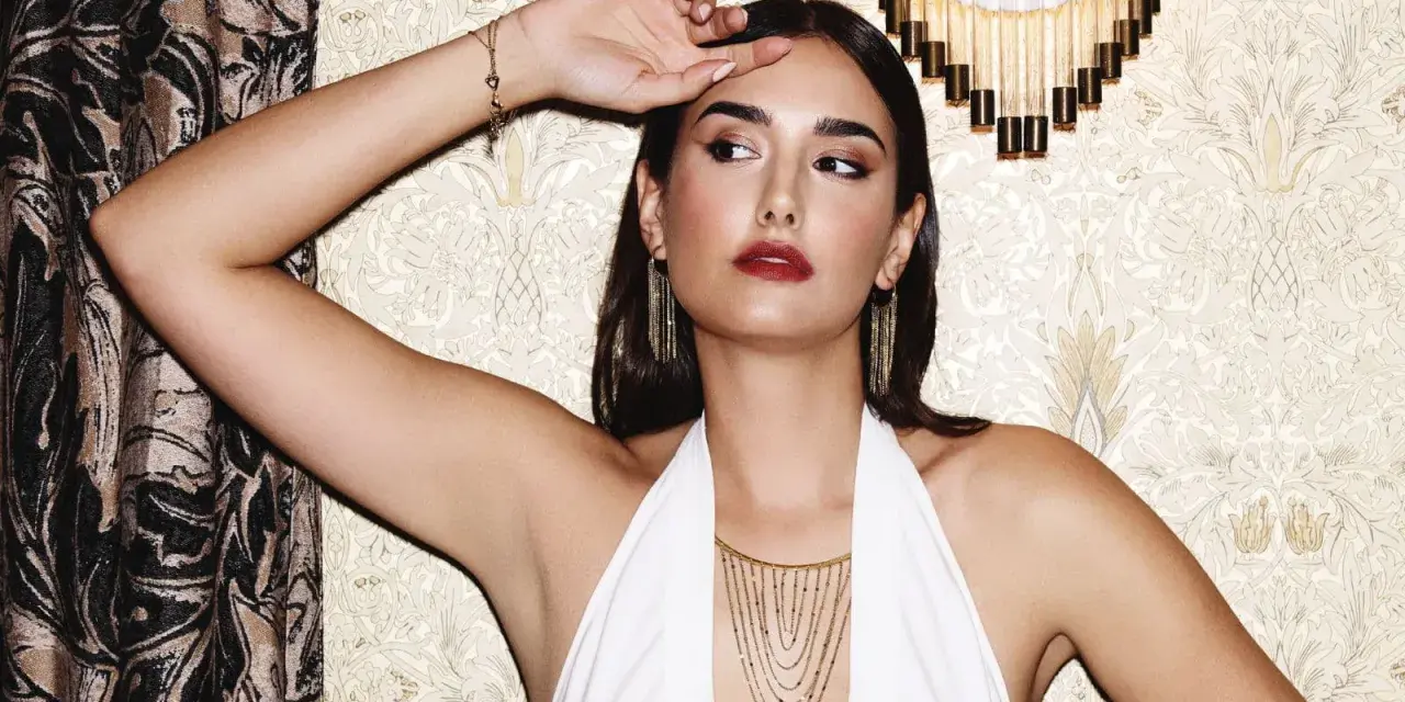 Mujer con pendientes media luna dorados, collar en cascada y pulsera de corazones, posando con un vestido blanco halter.