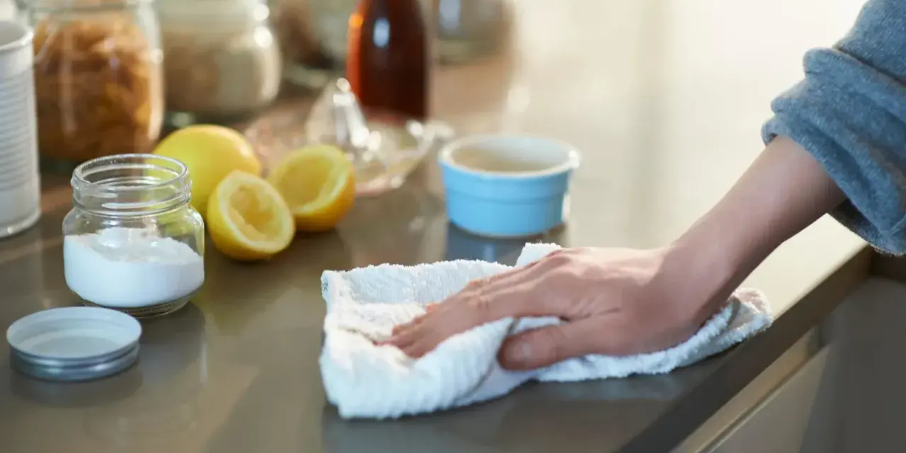 Mano limpiando una encimera con un paño blanco, junto a un frasco de bicarbonato y limones.