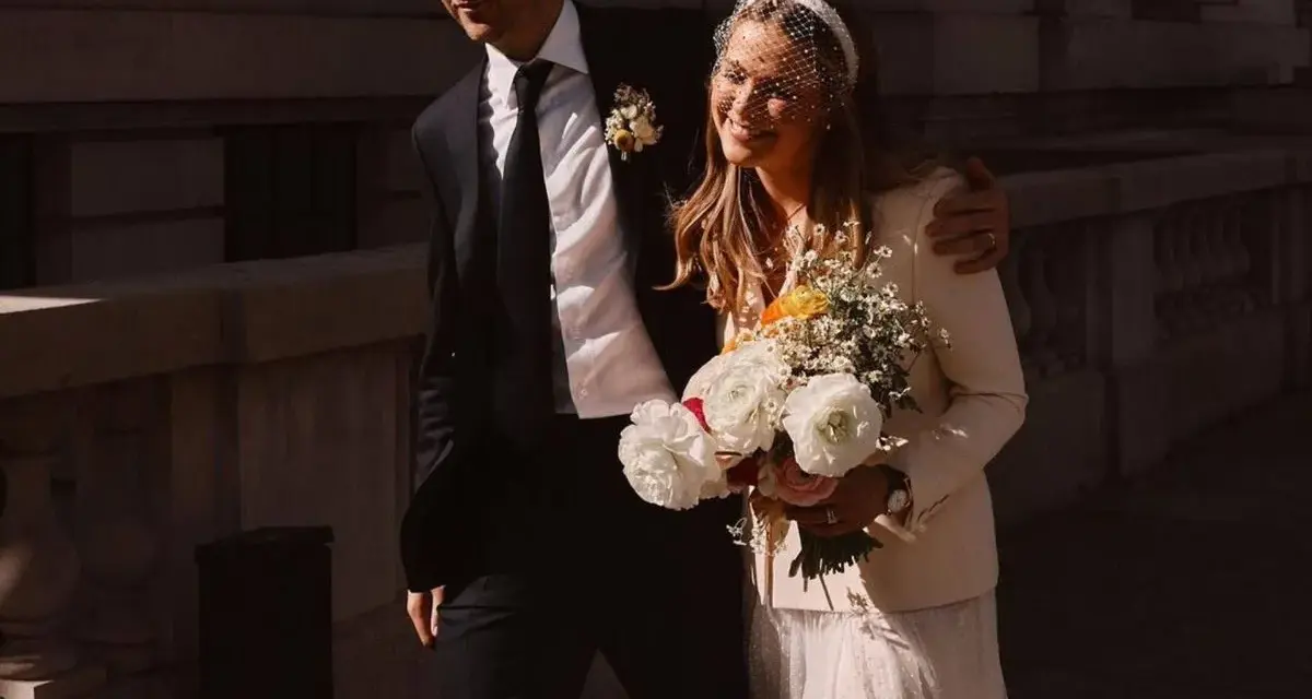 Novia sonriente con velo y ramo, luciendo una chaqueta novia boho sobre su vestido. El novio, con traje oscuro, la abraza.