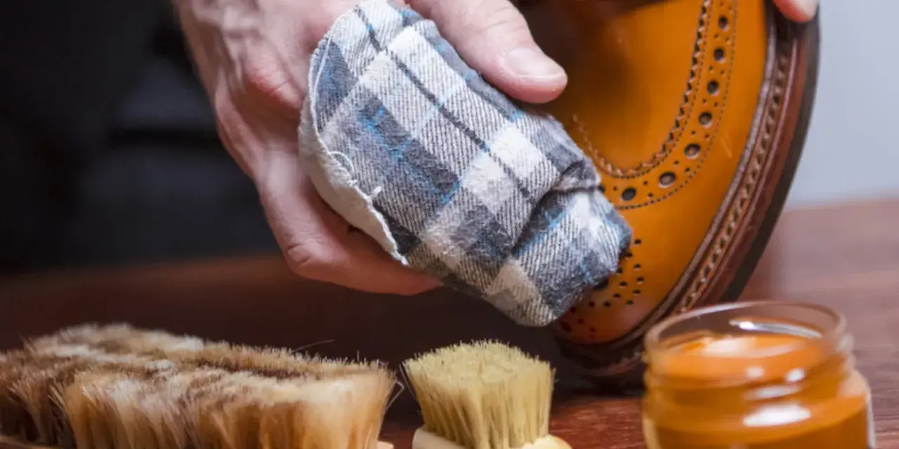 Mano limpiando un zapato de piel con un paño. Cerca, un cepillo y crema para zapatos. Aprende como limpiar zapatos de piel.