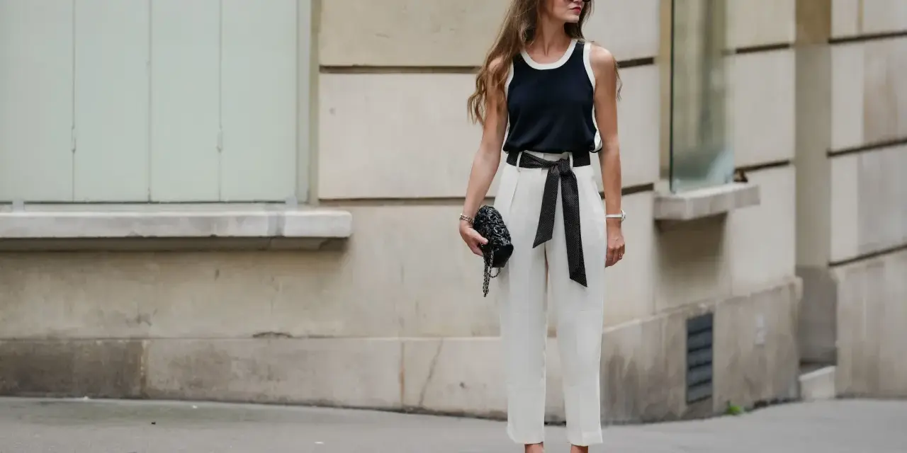 Mujer con elegante pantalón lino blanco, top negro y bolso de mano, posando con confianza.