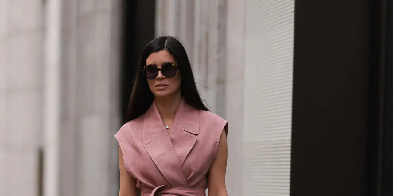 Mujer con gafas de sol y un elegante chaleco de lino rosa, atado a la cintura, pasea por la ciudad.