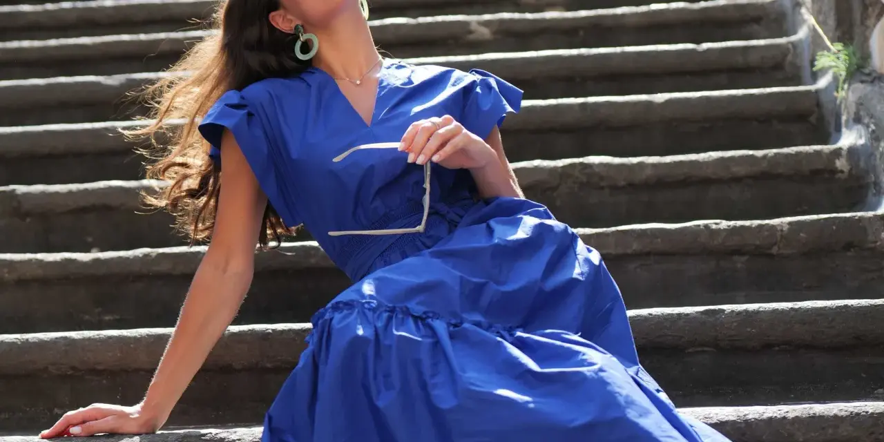Mujer con vestido azul marino, gafas de sol en mano, sentada en escalones de piedra. Perfecta para combinar con zapatos para vestido azul marino.