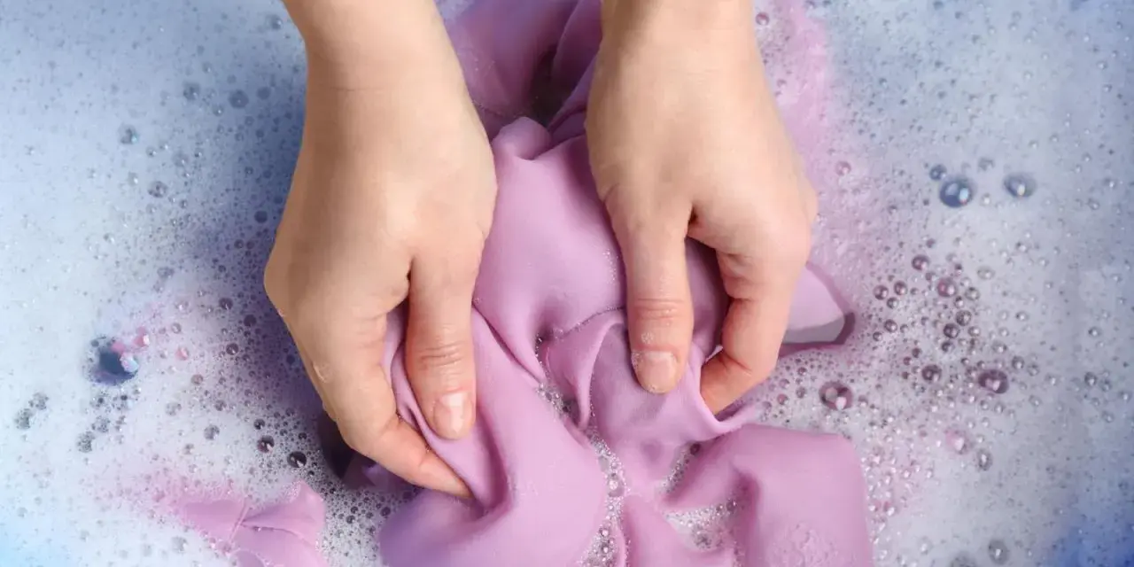 Manos lavando ropa morada a mano en agua con jabón y burbujas.