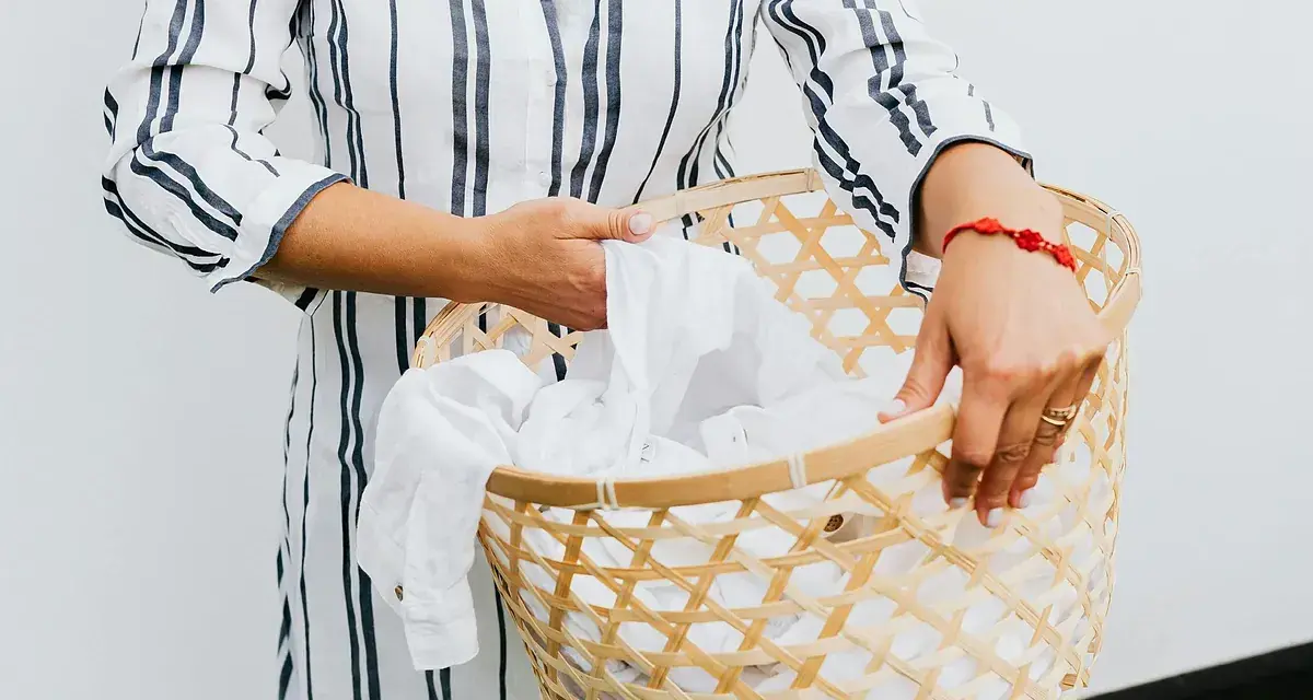 Manos sostienen una cesta de mimbre llena de ropa blanca, lista para lavar camiseta a mano.