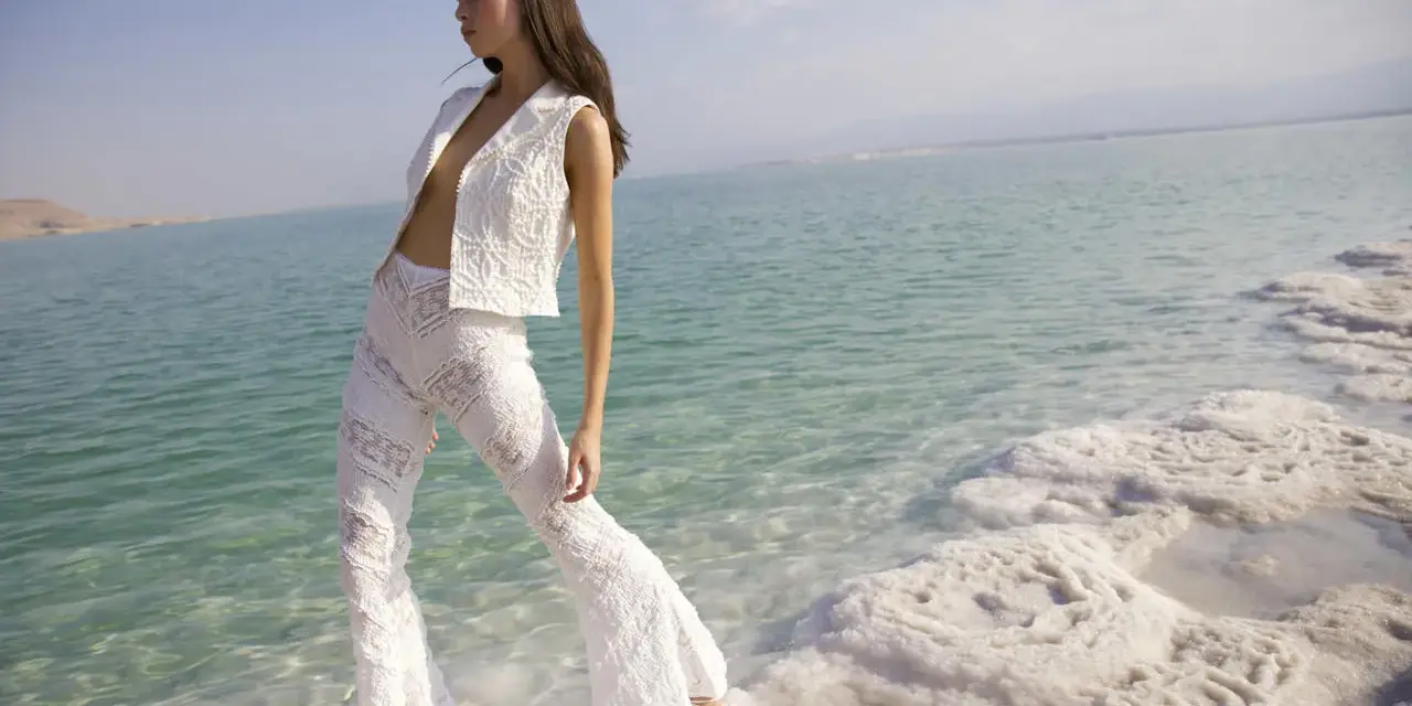 Mujer con traje blanco de encaje, posando en la orilla de un mar turquesa con formaciones de sal. Un look ibiza perfecto para el verano.
