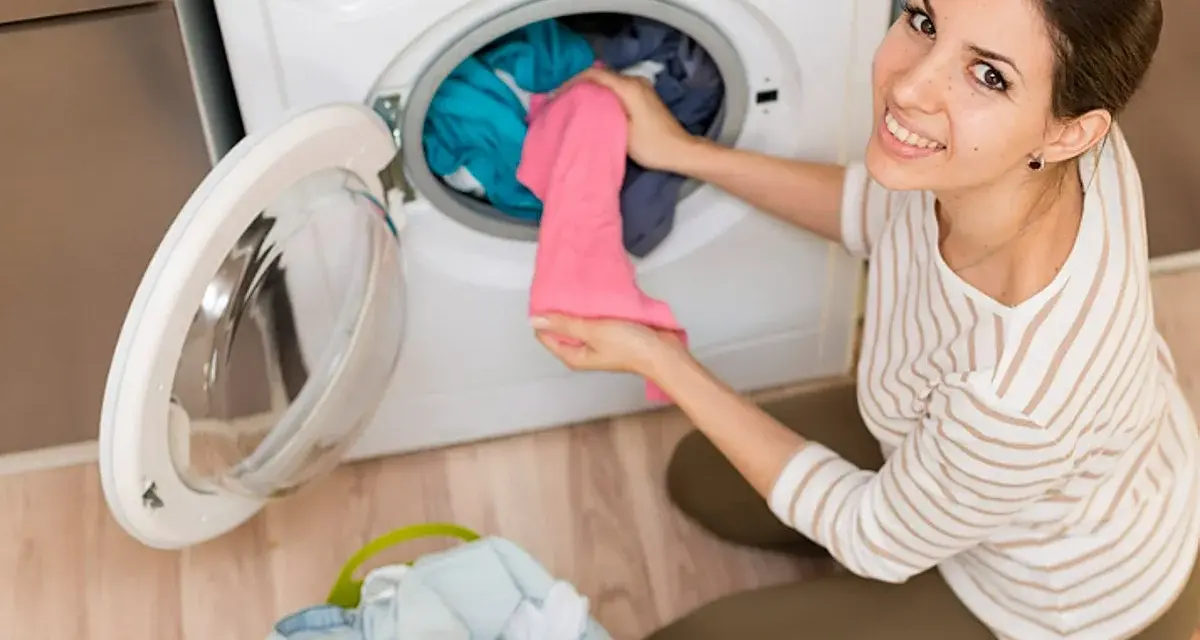 Mujer sonriente carga ropa en lavadora. El detergente sirve para dejar la ropa impecable y eliminar manchas.