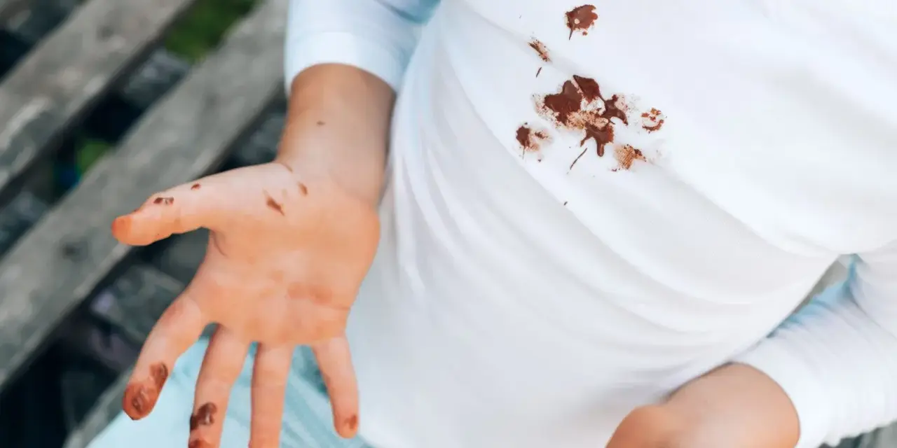 Manos y camiseta blanca con manchas de chocolate. Un niño muestra sus manos sucias y su ropa manchada, buscando cómo quitar manchas de chocolate ya lavadas.