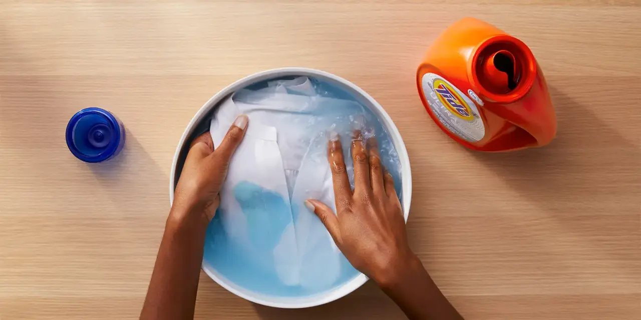 Manos lavando ropa blanca en un bol con agua y detergente. A un lado, un bote de detergente y un tapón azul.