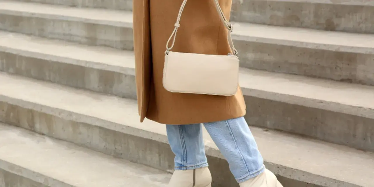 Mujer con abrigo camel, jeans y bolso crema, lista para combinar outfit con botas beige.