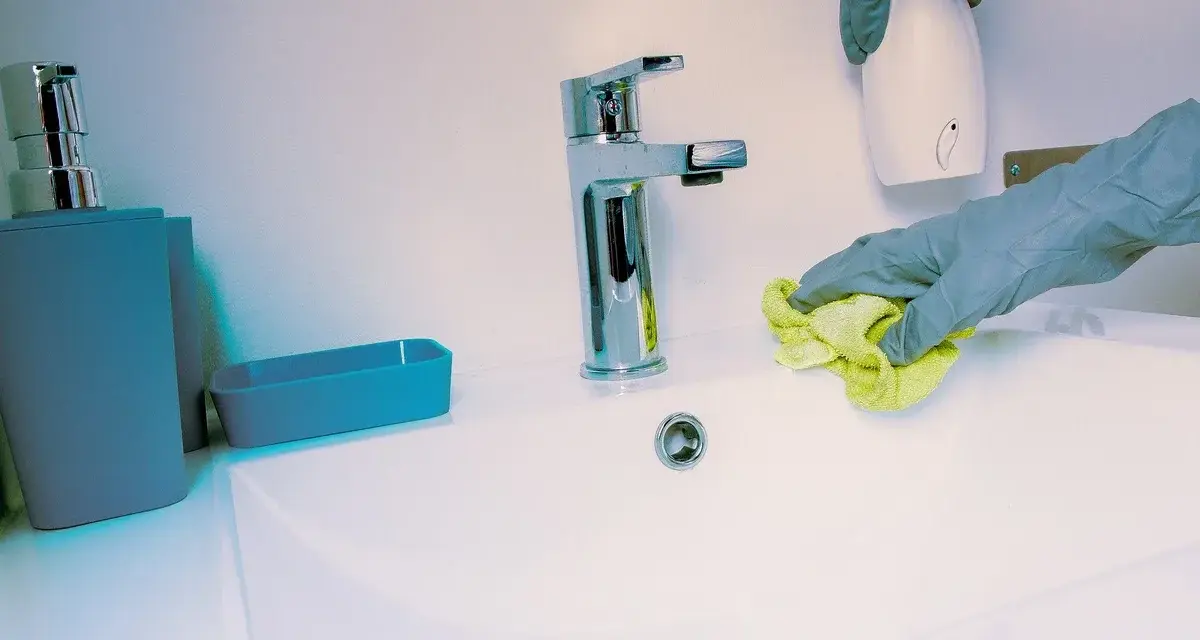 Manos con guantes limpian lavabo. Se usa un paño amarillo y un dispensador blanco.