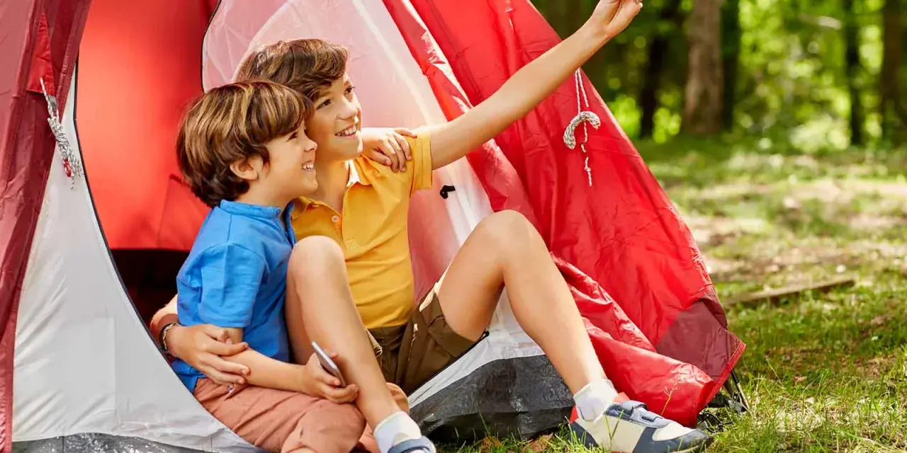 Dos niños, uno con talla niño 7 años, se toman un selfie sonrientes dentro de una tienda de campaña roja en un bosque.