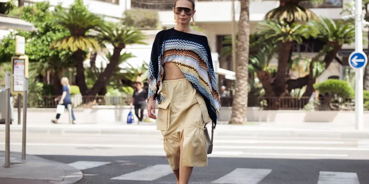 Mujer con un top corto de crochet y una falda vaporosa color beige, paseando por la calle con palmeras de fondo.