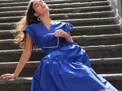 Mujer con vestido azul marino, gafas de sol en mano, sentada en escalones de piedra. Perfecta para combinar con zapatos para vestido azul marino.