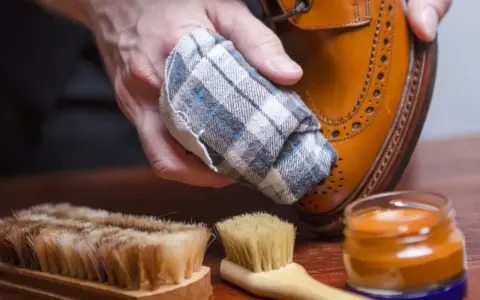 Mano limpiando un zapato de piel con un paño. Cerca, un cepillo y crema para zapatos. Aprende como limpiar zapatos de piel.