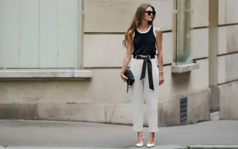 Mujer con elegante pantalón lino blanco, top negro y bolso de mano, posando con confianza.