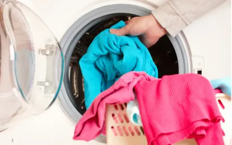 Mano cargando ropa azul en lavadora. Aprende como limpiar la lavadora para que tu ropa quede impecable.