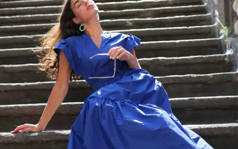 Mujer con vestido azul marino, gafas de sol en mano, sentada en escalones de piedra. Perfecta para combinar con zapatos para vestido azul marino.