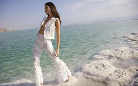 Mujer con traje blanco de encaje, posando en la orilla de un mar turquesa con formaciones de sal. Un look ibiza perfecto para el verano.