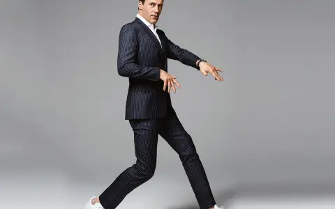 Hombre con traje a rayas y zapatillas deportivas blancas, en pose dinámica sobre fondo gris.