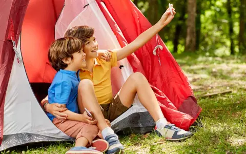 Dos niños, uno con talla niño 7 años, se toman un selfie sonrientes dentro de una tienda de campaña roja en un bosque.