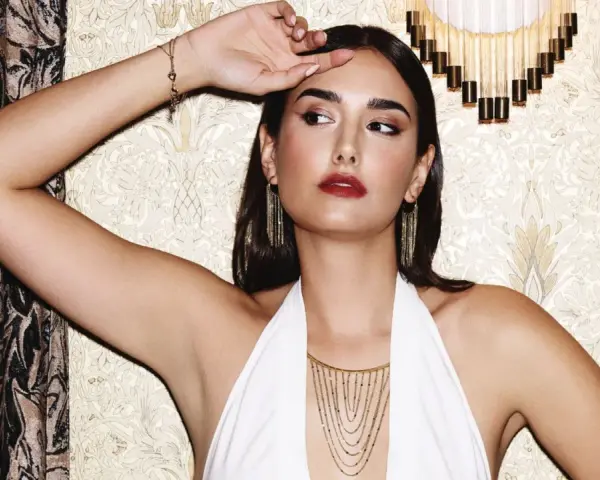 Mujer con pendientes media luna dorados, collar en cascada y pulsera de corazones, posando con un vestido blanco halter.