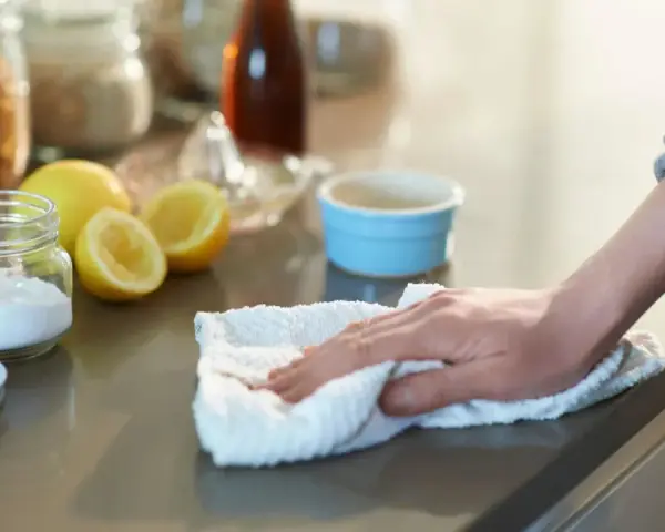 Mano limpiando una encimera con un paño blanco, junto a un frasco de bicarbonato y limones.