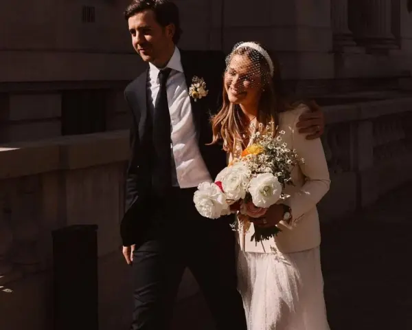 Novia sonriente con velo y ramo, luciendo una chaqueta novia boho sobre su vestido. El novio, con traje oscuro, la abraza.