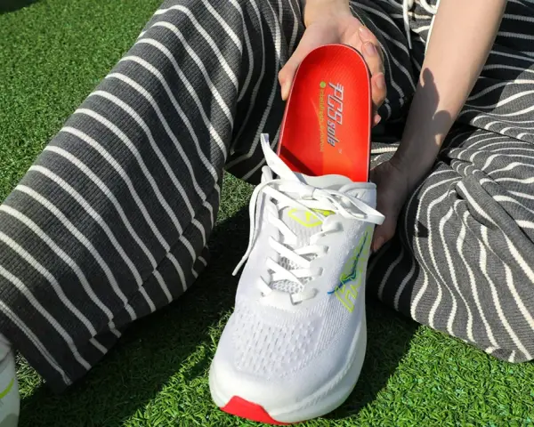 Persona muestra una plantilla roja para el arco plantar, lista para ser colocada en un zapato deportivo blanco.