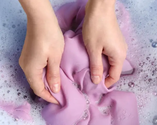Manos lavando ropa morada a mano en agua con jabón y burbujas.