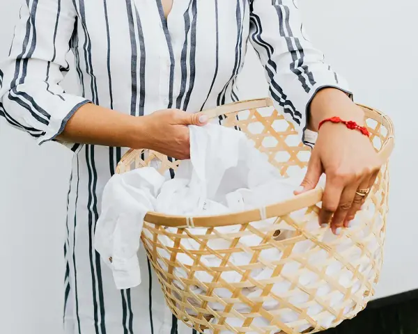 Manos sostienen una cesta de mimbre llena de ropa blanca, lista para lavar camiseta a mano.