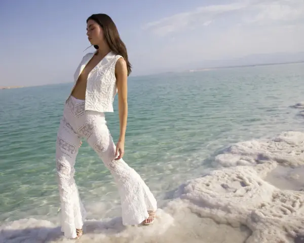 Mujer con traje blanco de encaje, posando en la orilla de un mar turquesa con formaciones de sal. Un look ibiza perfecto para el verano.