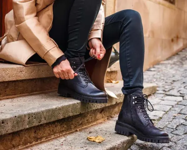 Persona se sienta en escalones de piedra, atándose unos pantalones anchos con botas militares negras.
