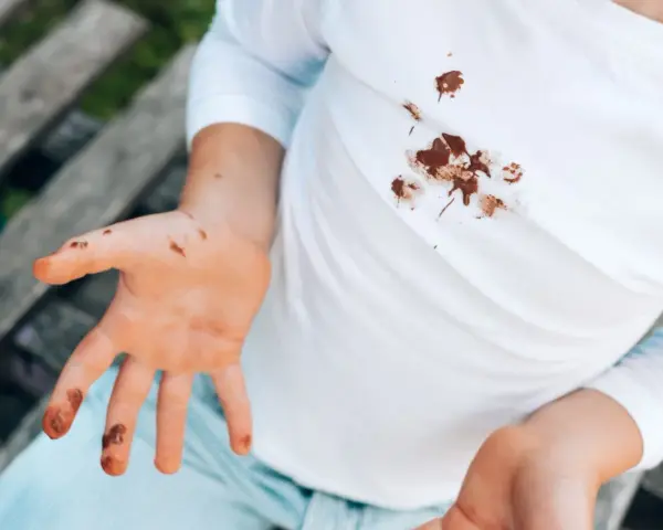 Manos y camiseta blanca con manchas de chocolate. Un niño muestra sus manos sucias y su ropa manchada, buscando cómo quitar manchas de chocolate ya lavadas.