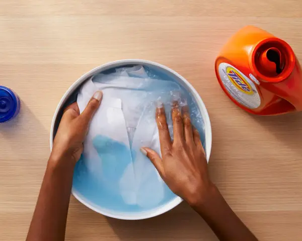 Manos lavando ropa blanca en un bol con agua y detergente. A un lado, un bote de detergente y un tapón azul.