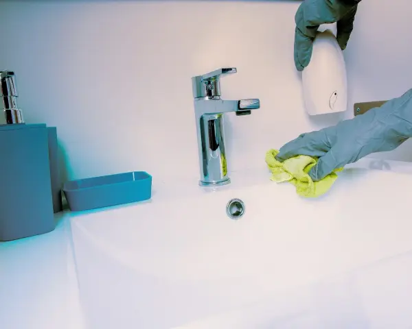 Manos con guantes limpian lavabo. Se usa un paño amarillo y un dispensador blanco.