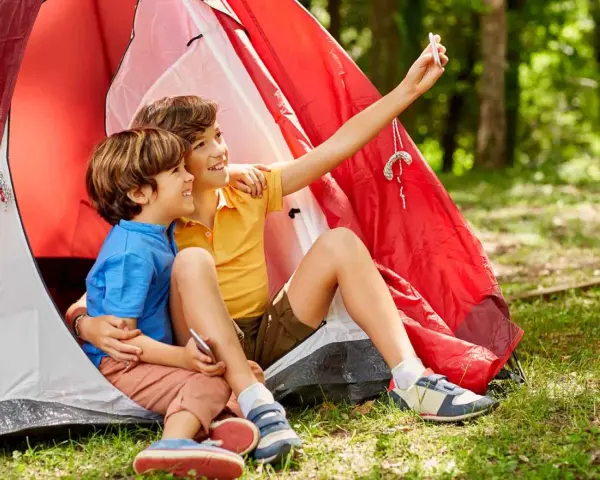Dos niños, uno con talla niño 7 años, se toman un selfie sonrientes dentro de una tienda de campaña roja en un bosque.