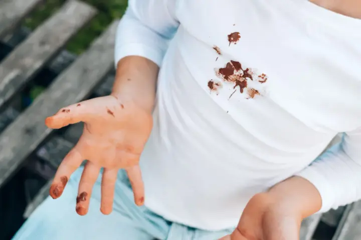 Manos y camiseta blanca con manchas de chocolate. Un niño muestra sus manos sucias y su ropa manchada, buscando cómo quitar manchas de chocolate ya lavadas.