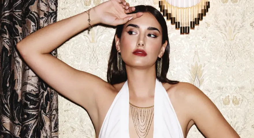 Mujer con pendientes media luna dorados, collar en cascada y pulsera de corazones, posando con un vestido blanco halter.