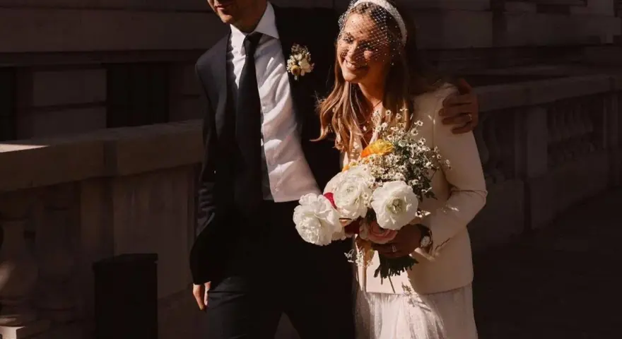 Novia sonriente con velo y ramo, luciendo una chaqueta novia boho sobre su vestido. El novio, con traje oscuro, la abraza.