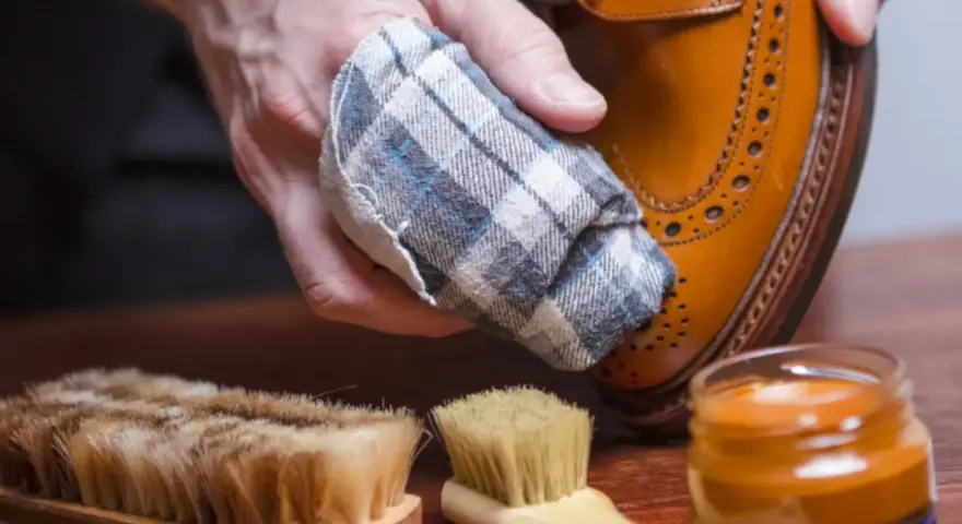 Mano limpiando un zapato de piel con un paño. Cerca, un cepillo y crema para zapatos. Aprende como limpiar zapatos de piel.
