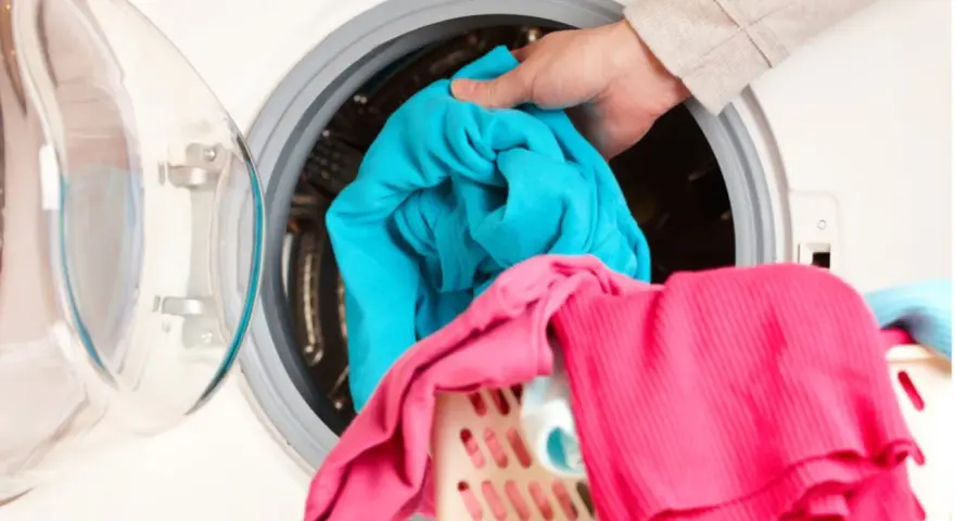 Mano cargando ropa azul en lavadora. Aprende como limpiar la lavadora para que tu ropa quede impecable.